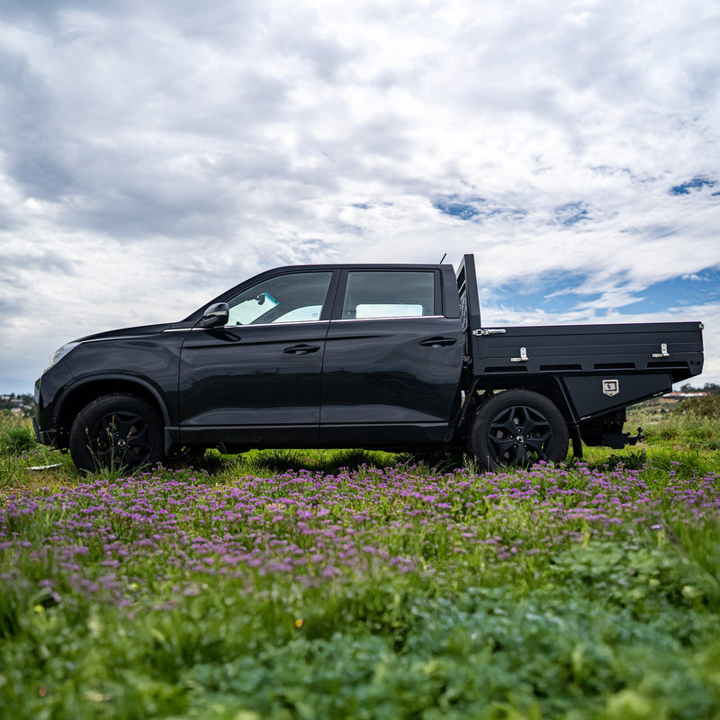 Load image into Gallery viewer, A black dual cab ute equipped with a sleek black Deluxe DMW Alloy Tray and under-tray toolboxes, parked in a vibrant field of wildflowers under a partly cloudy sky. This lightweight yet durable tray setup is ideal for both urban utility and off-road capability.