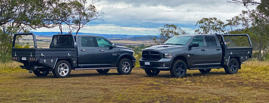 Two RAM 1500 dual cab utes, both equipped with custom black DMW XT Alloy Trays, are parked on a scenic hilltop with rolling farmland in the background. Each tray features under-tray toolboxes, tie-down points, and optional headboards, showcasing DMW's capability for American full-size ute builds.