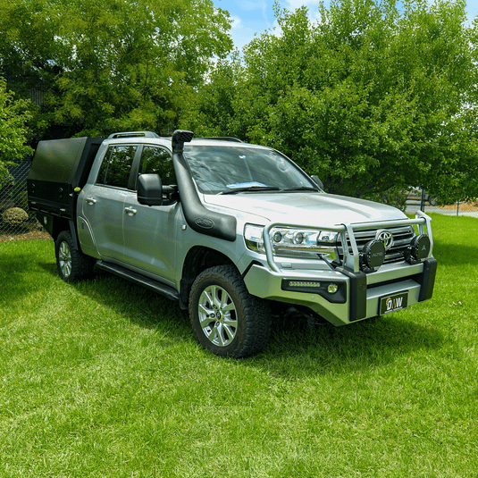 A silver Toyota LandCruiser 200 Series dual cab ute featuring a DMW Deluxe Alloy Tray with compact black canopy, photographed on green grass in a park setting. Outfitted with a snorkel, bull bar, and driving lights, this build is ideal for off-road touring with secure rear storage and protection.