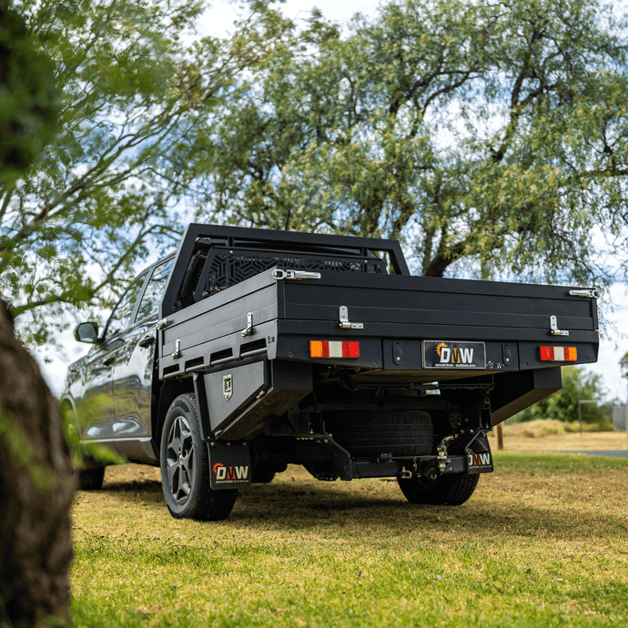 A sleek black DMW Deluxe Alloy Tray mounted on a dual cab ute, parked on green grass beneath shaded trees. The tray features integrated tie rails, combing rails, and a heavy-duty mesh headboard—offering a refined yet rugged solution for trade and recreational use.
