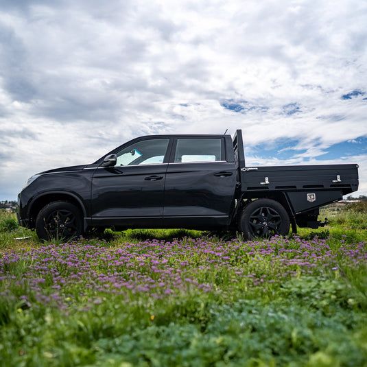 A black dual cab ute equipped with a sleek black Deluxe DMW Alloy Tray and under-tray toolboxes, parked in a vibrant field of wildflowers under a partly cloudy sky. This lightweight yet durable tray setup is ideal for both urban utility and off-road capability.