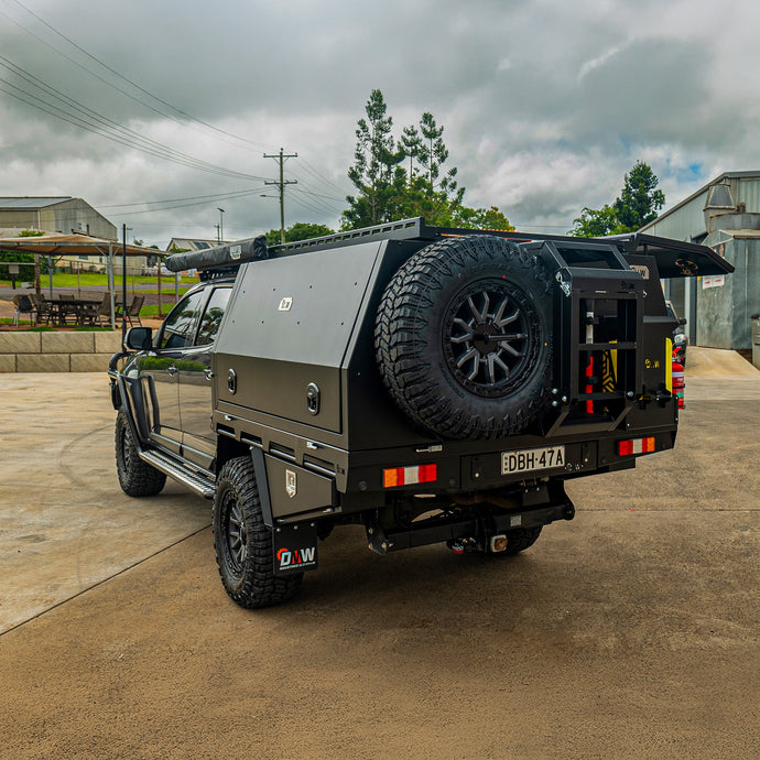 A black Y62 dual cab ute equipped with a DMW Deluxe Alloy Tray and enclosed canopy system featuring a rear spare wheel mount, fold down ladder, jerry can holder, and integrated under-tray toolboxes. Parked in an industrial lot, this build is purpose-designed for serious off-road touring and storage efficiency.