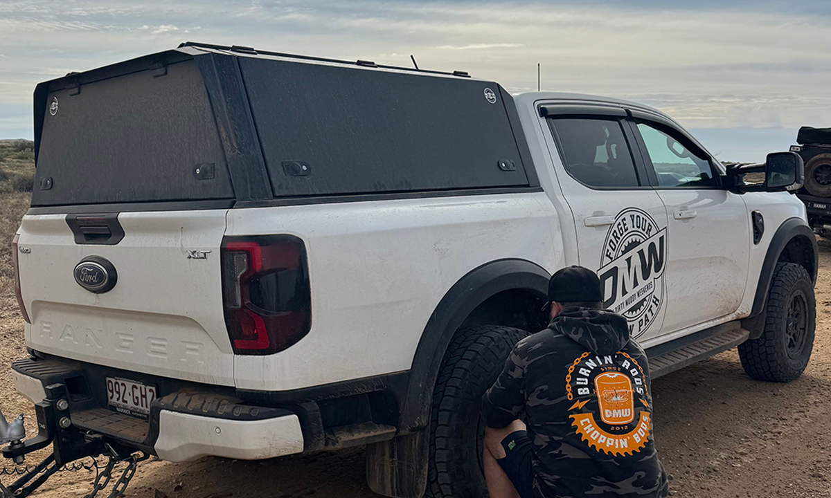 A white Ford Ranger XLT fitted with a DMW black tub replacement canopy, pictured in a dusty off-road environment with a team member attending to a tyre. The setup showcases durability and off-road readiness, making it ideal for overland adventures and rugged touring conditions.