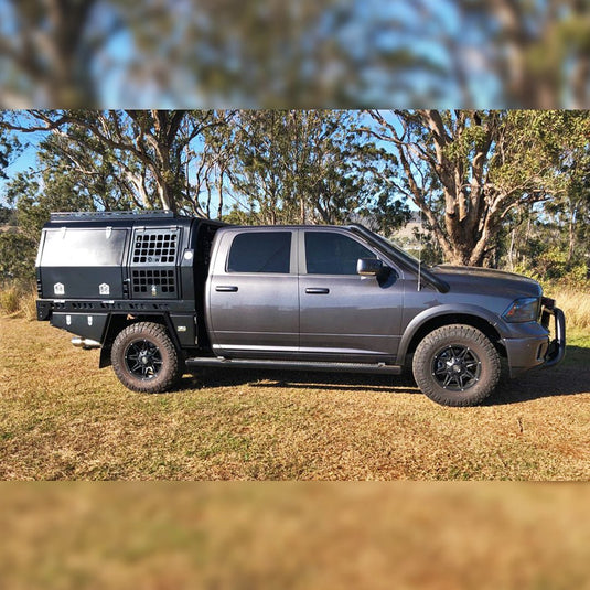 A grey RAM 1500 dual cab fitted with a black DMW Deluxe Alloy Tray and fully enclosed touring canopy featuring mesh side panels (Dog box) and dual access compartments. Parked on sunlit grass with bushland in the background, this American pickup showcases a rugged Australian tray and canopy conversion built for touring or work.