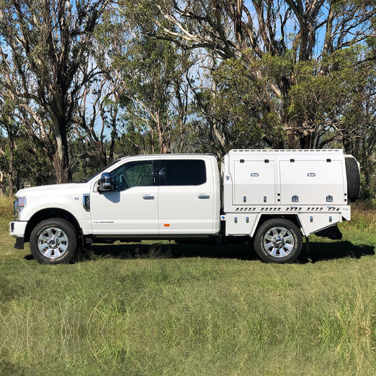 A white Ford F-250 Super Duty dual cab fitted with a DMW Deluxe Alloy Tray and full-length enclosed touring canopy, parked on green grass in a eucalyptus forest setting. The canopy features side access hatches, lockable compartments, and a rear spare wheel mount—engineered for long-haul off-road travel and maximum storage capacity.